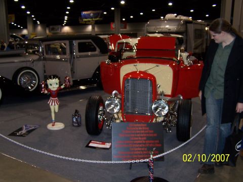 1979 Ford F-150 - A red hot rod is displayed at an indoor event, with a vintage car and a Betty Boop figure nearby.