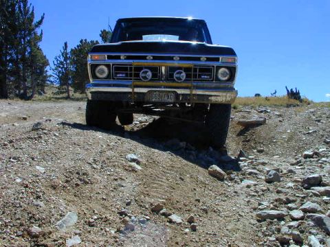 1979 Ford Bronco - Ground-level head-on view of a lifted dark Ford 4x4 straddling a rocky rutted trail, silhouetted against a clear blue sky with pines on the…