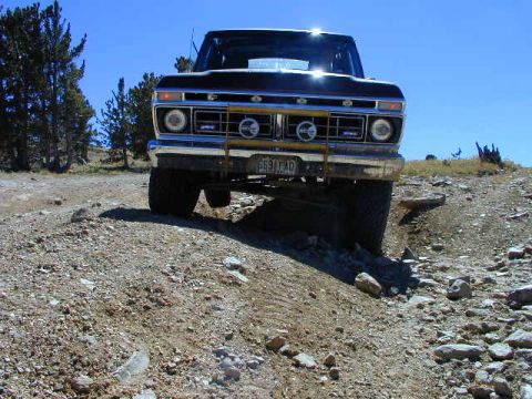 1979 Ford Bronco - Low ground-level front shot of a dark full-size Ford truck cresting a rocky dirt trail against a deep blue sky, with round headlights, a ch…