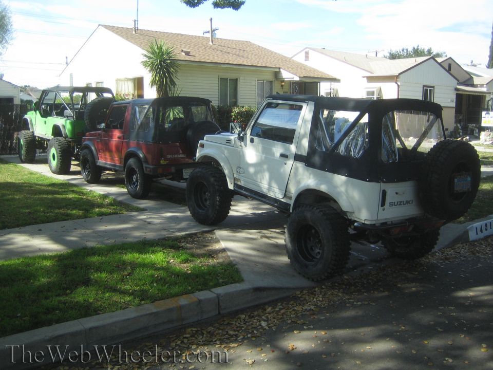 Three Suzuki Samurai SUVs parked in a row on a residential street. The closest is white, followed by red, then green