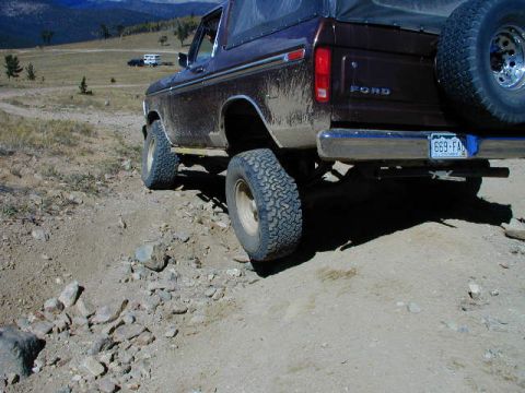 1979 Ford Bronco - Rear three-quarter view of a mud-splattered maroon Ford full-size Bronco flexing on a rocky mountain two-track, with FORD block lettering o…