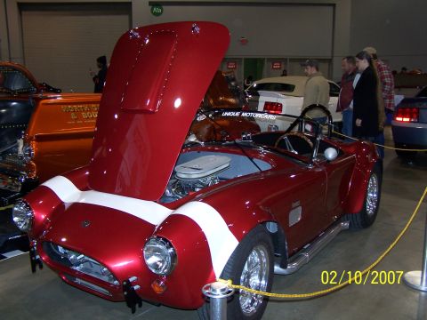 1979 Ford F-150 - A red AC Cobra replica is displayed with its hood open, revealing the engine. Other vehicles and people are visible in the background.