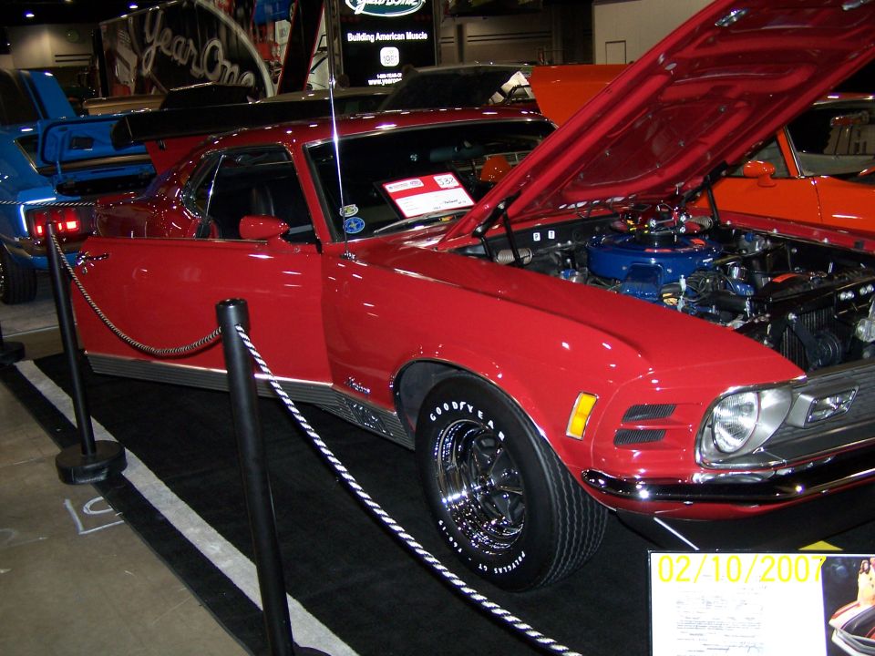 Red 1970 Ford Mustang Mach 1 with hood open, showing engine. Another blue car is visible to the left