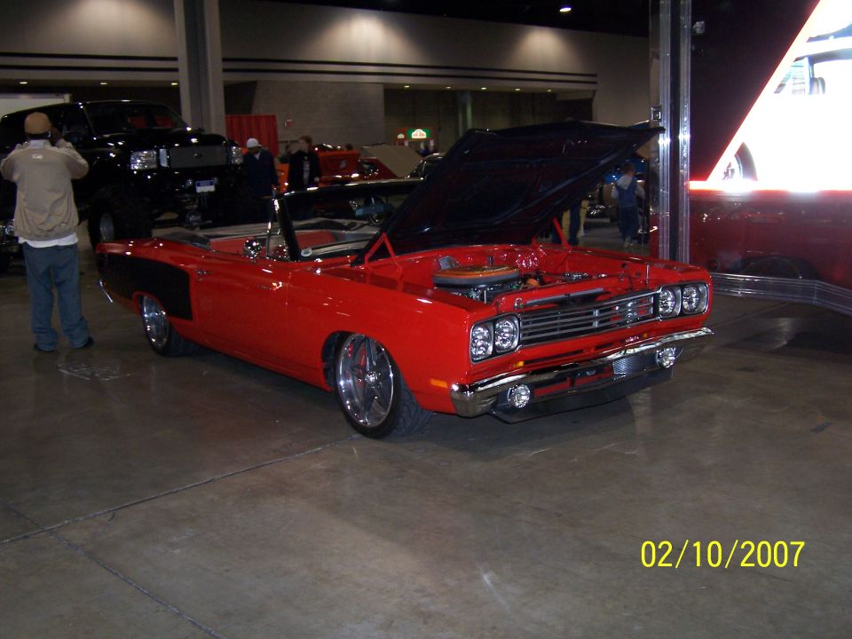 Red 1969 Plymouth Road Runner convertible with hood open, parked indoors. Black stripe on rear quarter panel. Chrome wheels