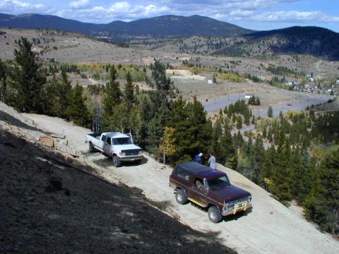 1979 Ford Bronco - A maroon 1979 Ford Bronco and a white pickup truck navigate a dirt road in a scenic, mountainous terrain.
