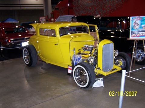 1979 Ford F-150 - A yellow hot rod coupe is displayed indoors with other vehicles and signage.