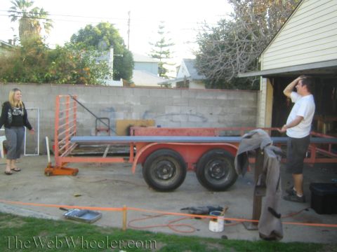 1987 Suzuki Samurai - A trailer carrying long metal pipes is parked in a yard with two people nearby.