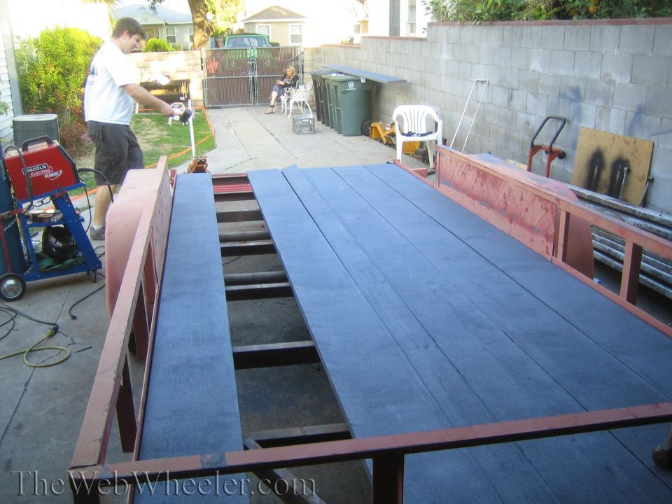 Red trailer with dark blue wooden planks, a man welding in the background