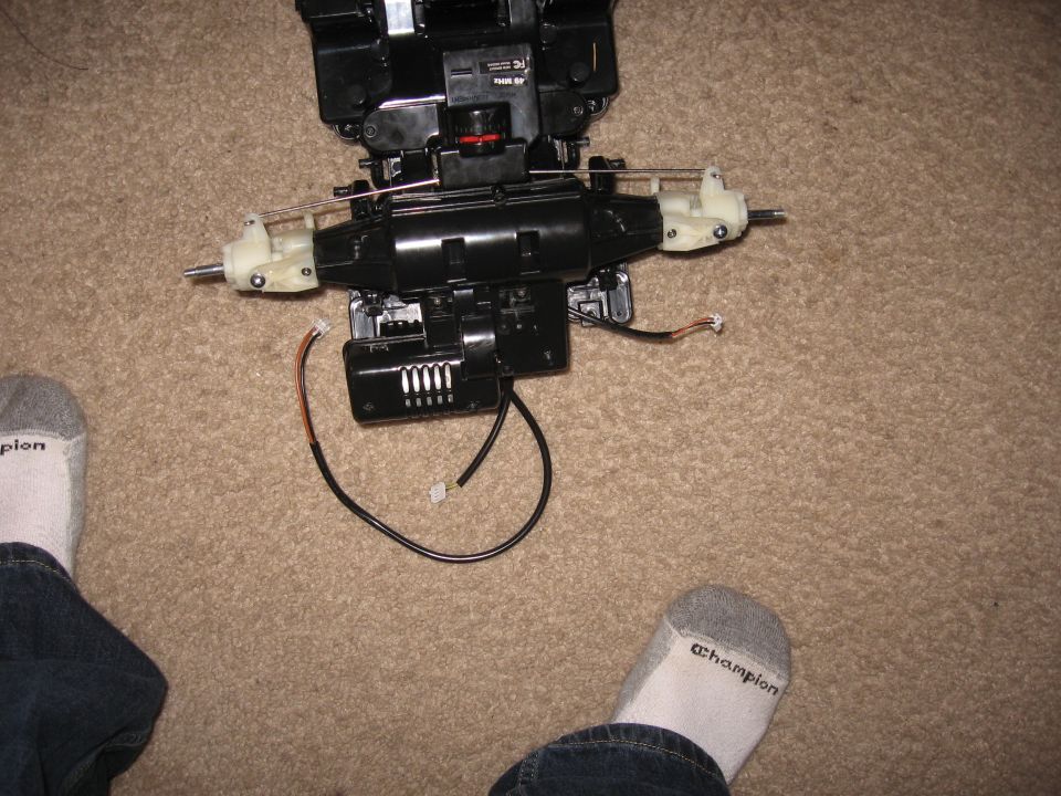 Remote control car chassis with suspension, motor, and wiring on carpet. Feet in socks visible
