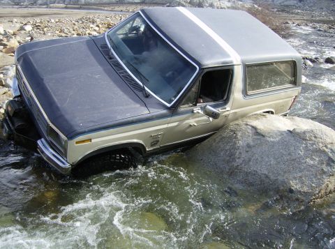 1986 Ford Bronco - This Ford Bronco is navigating a rocky riverbed, with water flowing around the tires and undercarriage.