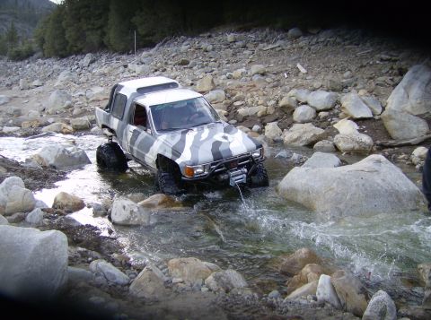 1986 Ford Bronco - This off-road truck, painted in a camouflage pattern, is shown carefully maneuvering through a shallow, rocky stream bed.