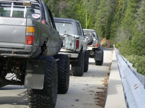 1986 Ford Bronco - Several heavily modified trucks drive in a convoy on a road through a wooded area.