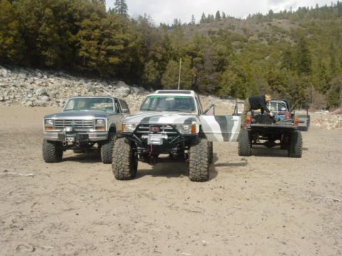 1986 Ford Bronco - Three modified trucks are parked on a sandy area with trees and hills in the background.