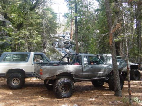 1986 Ford Bronco - Two modified trucks are parked off-road in a forest, with one appearing to be a pickup truck and the other an SUV.