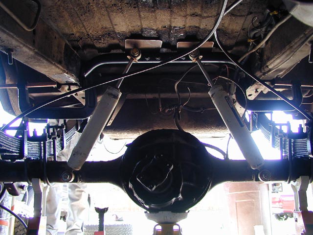 Underside view of a vehicle's rear axle, leaf springs, and shock absorbers. The vehicle is on jack stands