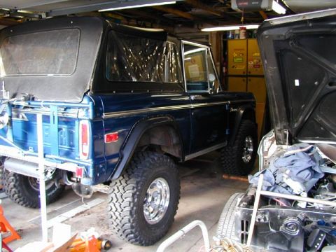 1979 Ford Bronco - Rear three-quarter view of a blue early Ford Bronco with a black soft top parked inside a home garage, showing the tailgate, cut rear fende…