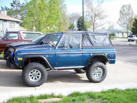 1979 Ford Bronco - Passenger-side profile of a blue early Ford Bronco with a black soft top, cursive Bronco fender script and black cut/flared fenders, parked…