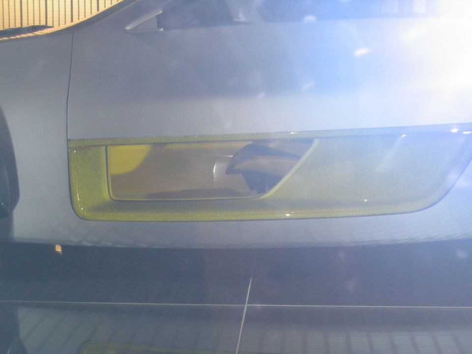 Silver car's front passenger side headlight assembly with yellow trim, under bright lights