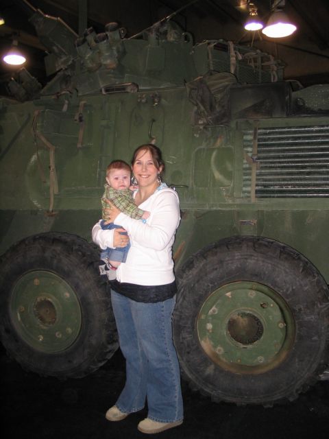 1993 Ford Taurus - A woman holds a baby in front of a large green military vehicle, likely on display at an auto show.