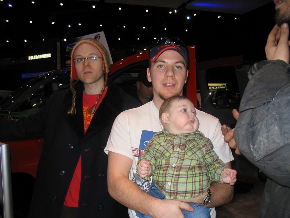 Three people and a baby pose in front of a red vehicle at an auto show with a "HUMMER" sign in the background