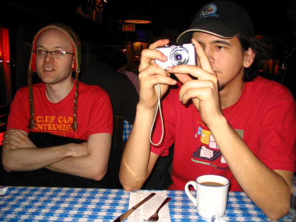 Man in red shirt with braided hat takes photo with digital camera; another man in red shirt sits with arms crossed