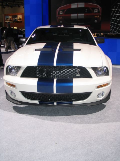 1993 Ford Taurus - A white 2007 Ford Mustang Shelby GT500 with blue racing stripes is displayed at an auto show.