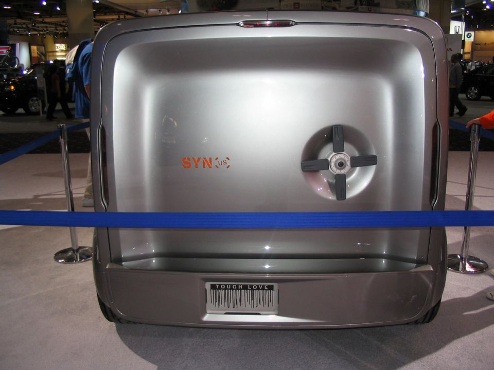 Rear view of a silver concept vehicle with "SYN:us" badging and a "TOUGH LOVE" license plate