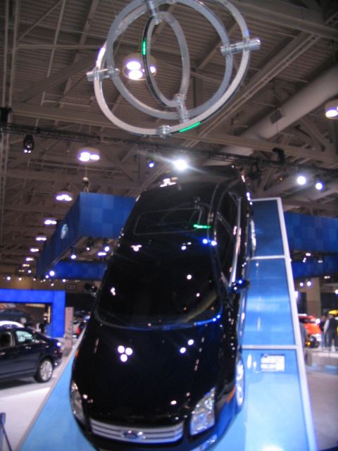 1993 Ford Taurus - A black sedan is presented on a ramp at an auto show, with a large, abstract metal sculpture hanging above.