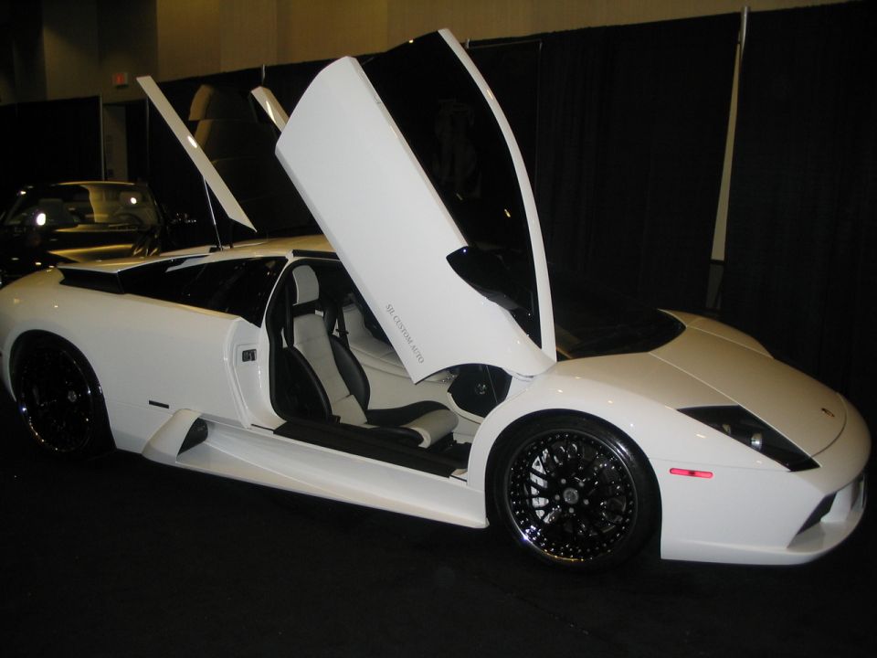 White sports car with black rims and scissor doors open, interior visible. Text 'SUPERMOTORS' at bottom right