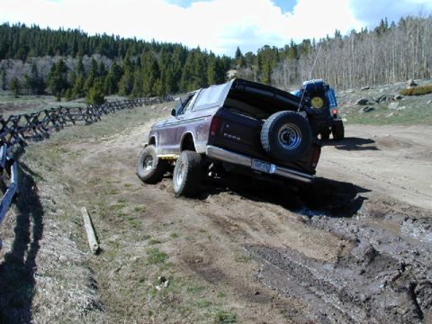1979 Ford Bronco - Maroon Ford full-size Bronco articulating through a muddy rut beside a split-rail fence on a forest trail, viewed from behind with FORD tai…