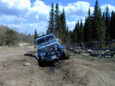 1979 Ford Bronco - Blue Jeep CJ dropping a front wheel into a deep mud hole on a forest two-track, front three-quarter view with a winch bumper, seven-slot gr…