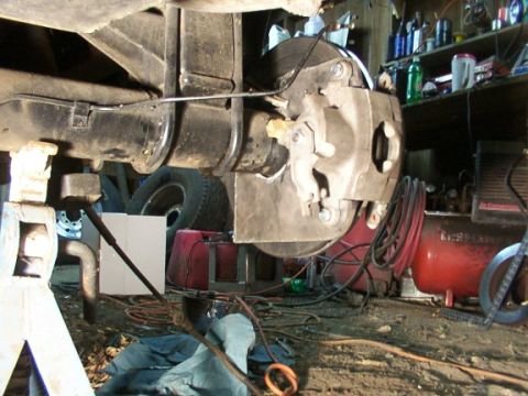 1995 Ford C-350 (Centurion Conversions) - Close-up view of a vehicle's rear axle and brake assembly, supported by jack stands in a cluttered workshop environment.