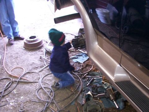 1995 Ford C-350 (Centurion Conversions) - A child is working on the brake assembly of a vehicle in a garage with tools and parts around them.