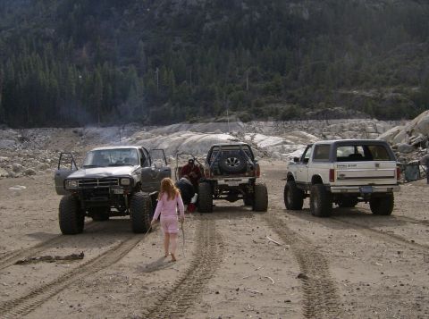1986 Ford Bronco - Three modified off-road vehicles are parked on a dirt lot, with a young girl in pink pajamas walking between them.
