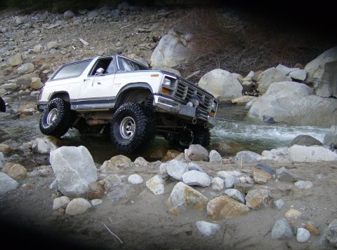 1986 Ford Bronco - A white 1980s Ford Bronco tackles a challenging off-road course, driving through a shallow, rocky stream.