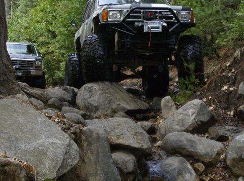 1986 Ford Bronco - Two modified 4x4s navigate a challenging rocky trail with dense foliage in the background.
