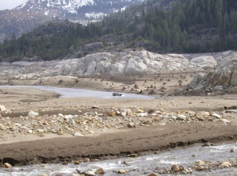1986 Ford Bronco - A vehicle navigates a dry, rocky riverbed with a mountainous, forested backdrop.