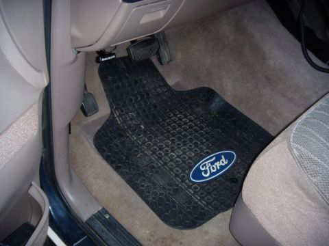 1992 Ford Bronco - The interior of a vehicle shows a black Ford-branded floor mat and the brake and gas pedals.