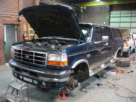 1995 Ford C-350 (Centurion Conversions) - A blue Ford SUV is undergoing maintenance with its hood open and front wheels removed, parked in a garage.