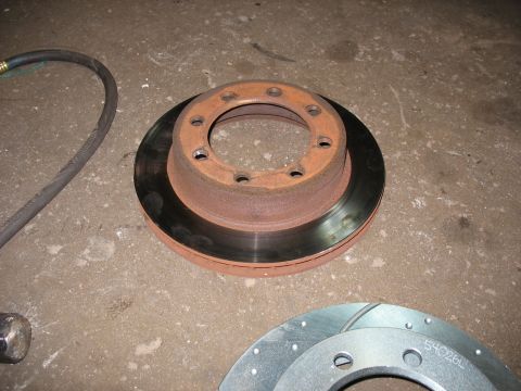 1995 Ford C-350 (Centurion Conversions) - A rusty brake rotor sits on the ground next to a new slotted brake rotor, with a hose and hammer visible.