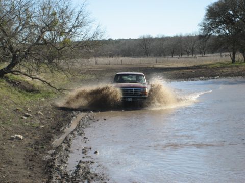 1986 Ford Bronco - A red pickup truck navigates a muddy water crossing, sending water and mud flying.