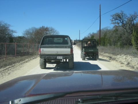 1986 Ford Bronco - A lifted Ford Bronco and a Kawasaki Mule drive down a dusty rural road under a clear blue sky.