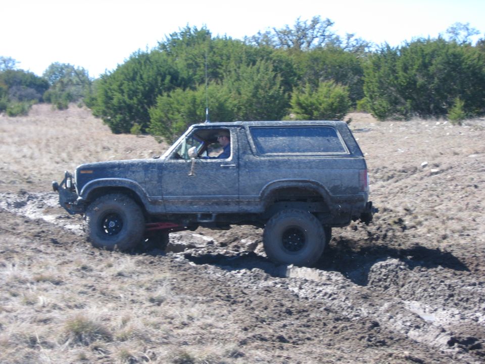 Mud-covered 1980s Ford Bronco driving through a muddy trail with trees in the background