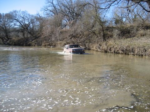 1996 Ford Bronco - A tan Ford Bronco navigates a shallow river crossing surrounded by bare trees and dry vegetation.