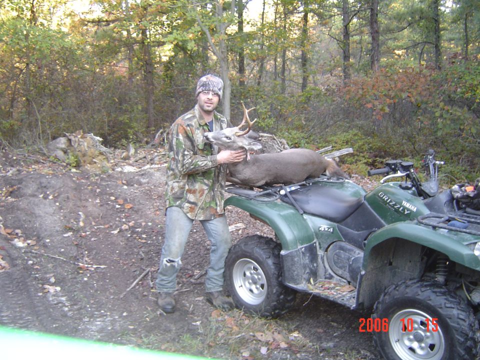 Hunter with a whitetail buck loaded on the rack of a green Yamaha Grizzly 4x4 ATV on a wooded dirt trail