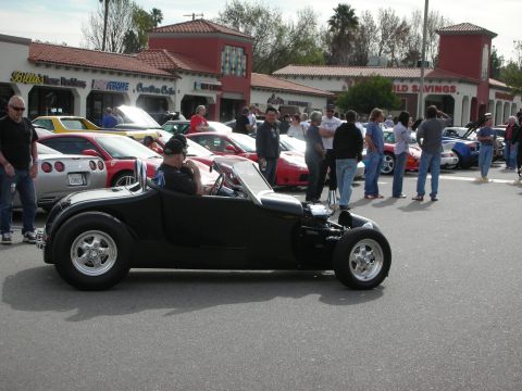 1998 Ford Crown Victoria - black low-slung hot rod roadster sits at a car show surrounded by spectators and parked Corvettes outside a strip mall.