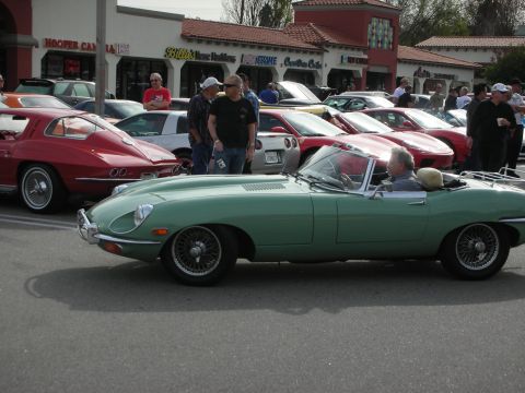 1998 Ford Crown Victoria - pale-green Jaguar E-Type roadster with wire wheels is driven through a car-show lot lined with classic cars and onlookers.