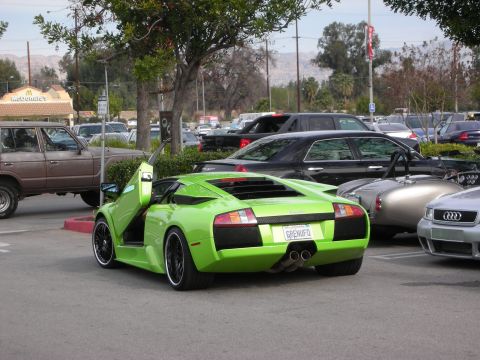 1998 Ford Crown Victoria - lime-green Lamborghini Murcielago parked with one scissor door raised, surrounded by other cars and distant hills.
