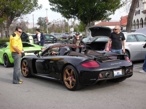 1998 Ford Crown Victoria - black Porsche Carrera GT with bronze wheels parked at a car show as a man in a yellow shirt looks on.