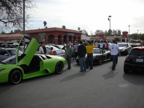 1998 Ford Crown Victoria - busy car-show lot with a green Lamborghini, doors raised, and a black Porsche surrounded by spectators outside a depot building.